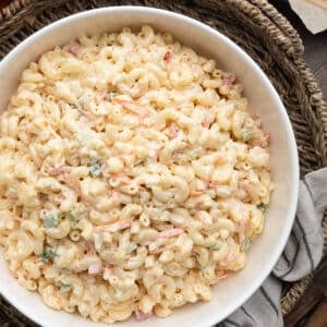 Close-up of creamy macaroni salad in a white serving bowl, featuring elbow macaroni coated in a mayonnaise-based dressing with shredded carrots, diced celery, onion, and bell peppers. The bowl sits on a woven tray with a striped kitchen towel beside it on a dark wood tabletop.
