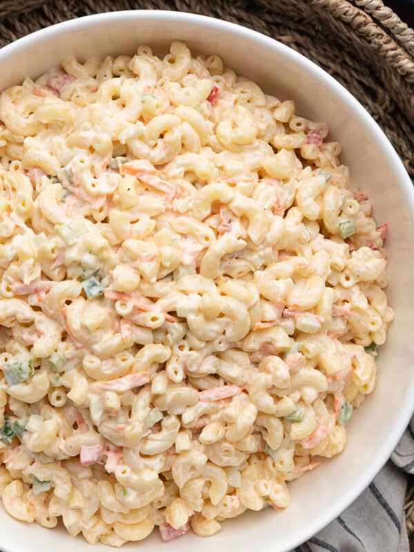 Close-up of creamy macaroni salad in a white serving bowl, featuring elbow macaroni coated in a mayonnaise-based dressing with shredded carrots, diced celery, onion, and bell peppers. The bowl sits on a woven tray with a striped kitchen towel beside it on a dark wood tabletop.