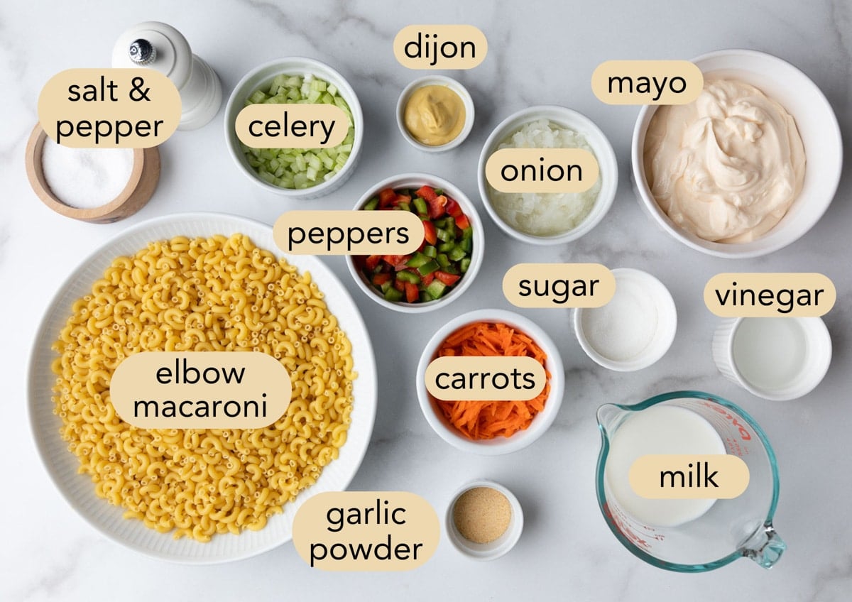 Overhead ingredient shot showing all the components needed to make creamy macaroni salad, including cooked elbow macaroni, mayonnaise, milk, celery, onion, bell peppers, shredded carrots, Dijon mustard, vinegar, sugar, garlic powder, salt, and pepper. Each ingredient is arranged in individual bowls and labeled on a white marble surface.