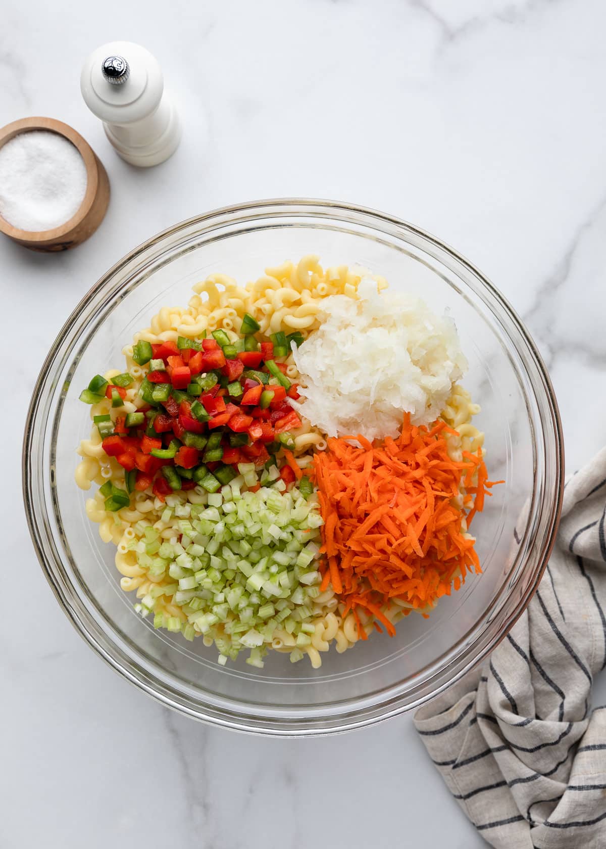 Cooked elbow macaroni topped with grated carrot, finely diced celery, minced onion, and diced red and green bell peppers, ready to be mixed into classic macaroni salad.