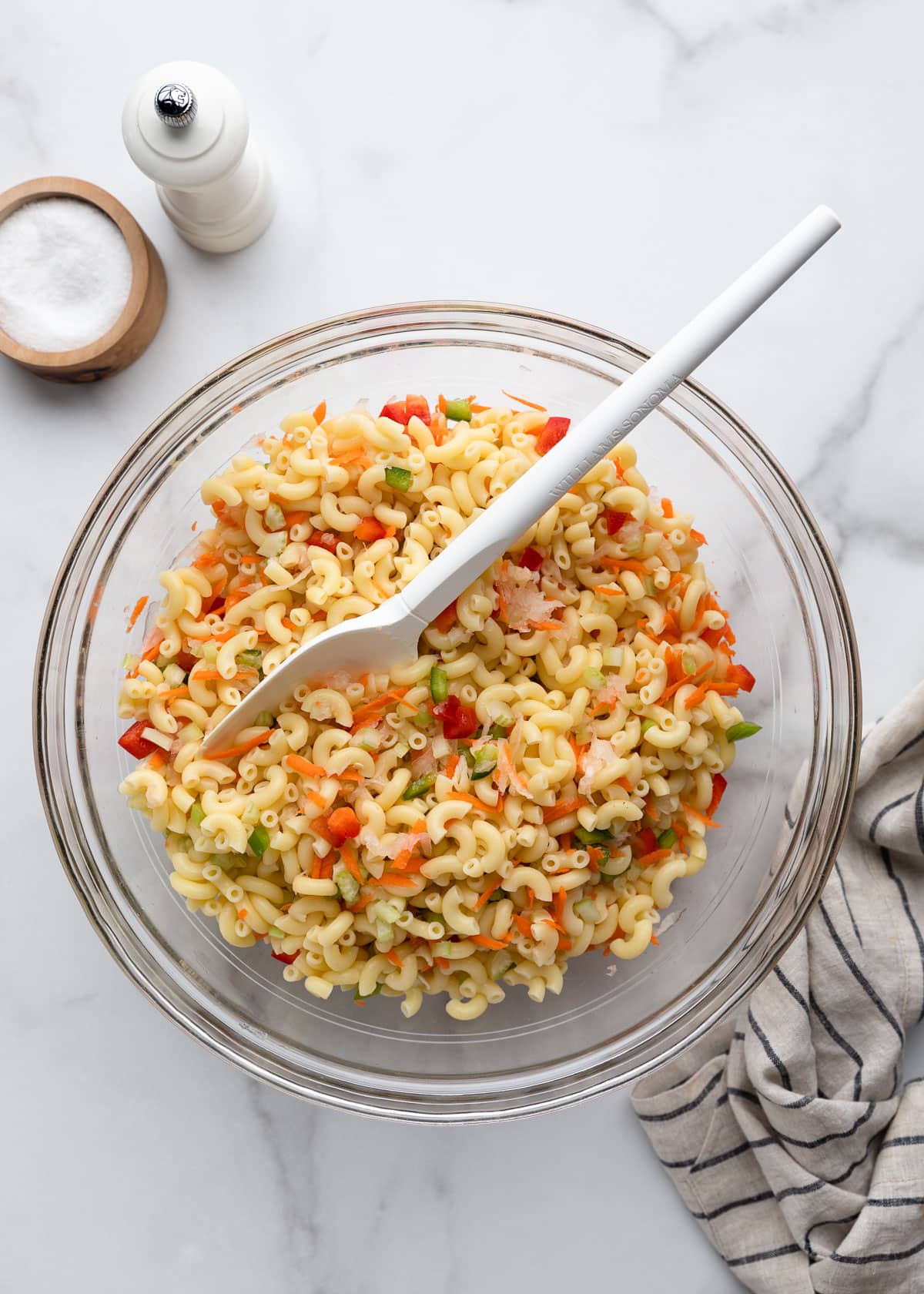 Macaroni salad tossed with shredded carrots and onion, finely diced celery, and red and green bell peppers in a large glass mixing bowl. A white serving spoon rests in the salad, with a salt cellar, pepper mill, and striped kitchen towel arranged nearby on a white marble surface.