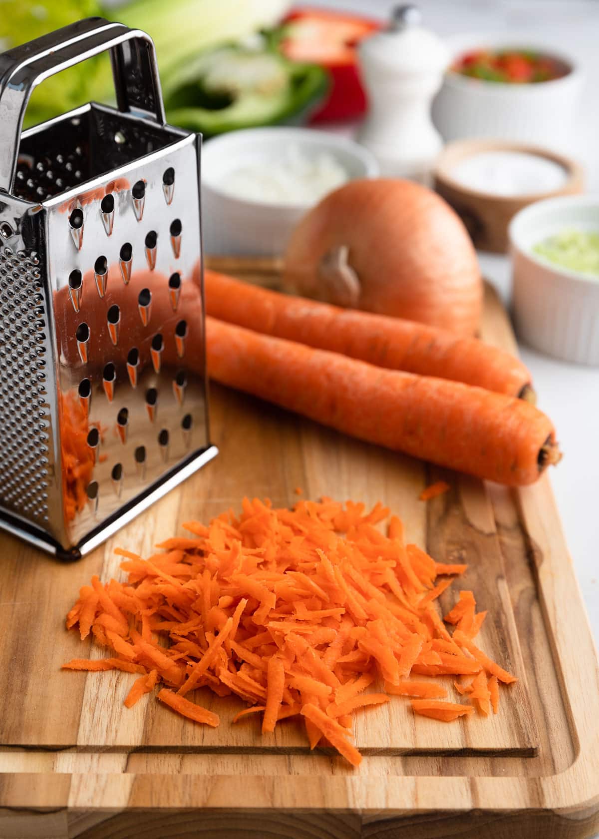 Box grater and freshly grated carrots on a wooden cutting board, with whole carrots, onion, celery, and other macaroni salad ingredients arranged in the background.