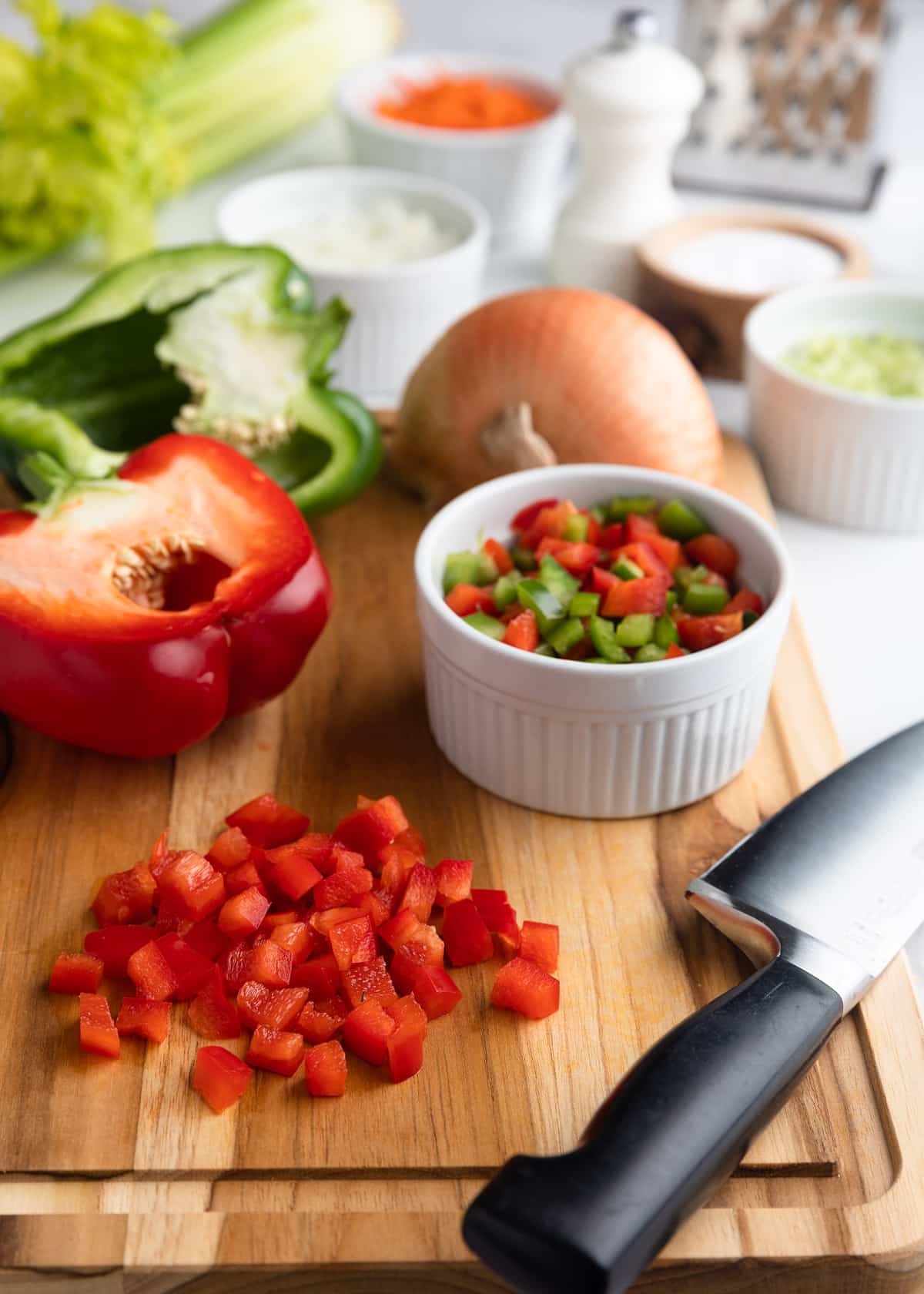 Diced bell peppers on a wooden cutting board beside a chef’s knife, with additional chopped vegetables and seasonings prepared for homemade macaroni salad.