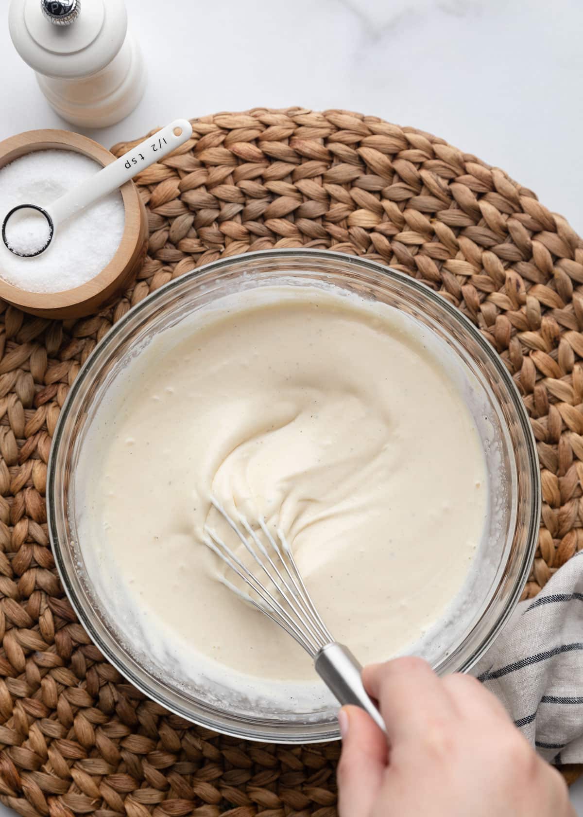 Creamy macaroni salad dressing being whisked in a glass mixing bowl, with a salt cellar and pepper mill nearby on a woven placemat.