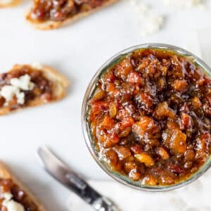 Close-up overhead view of a small glass bowl filled with chunky peach bacon jam, with blurred crostini topped with the jam and crumbled goat cheese, plus a spreader knife, in the background.