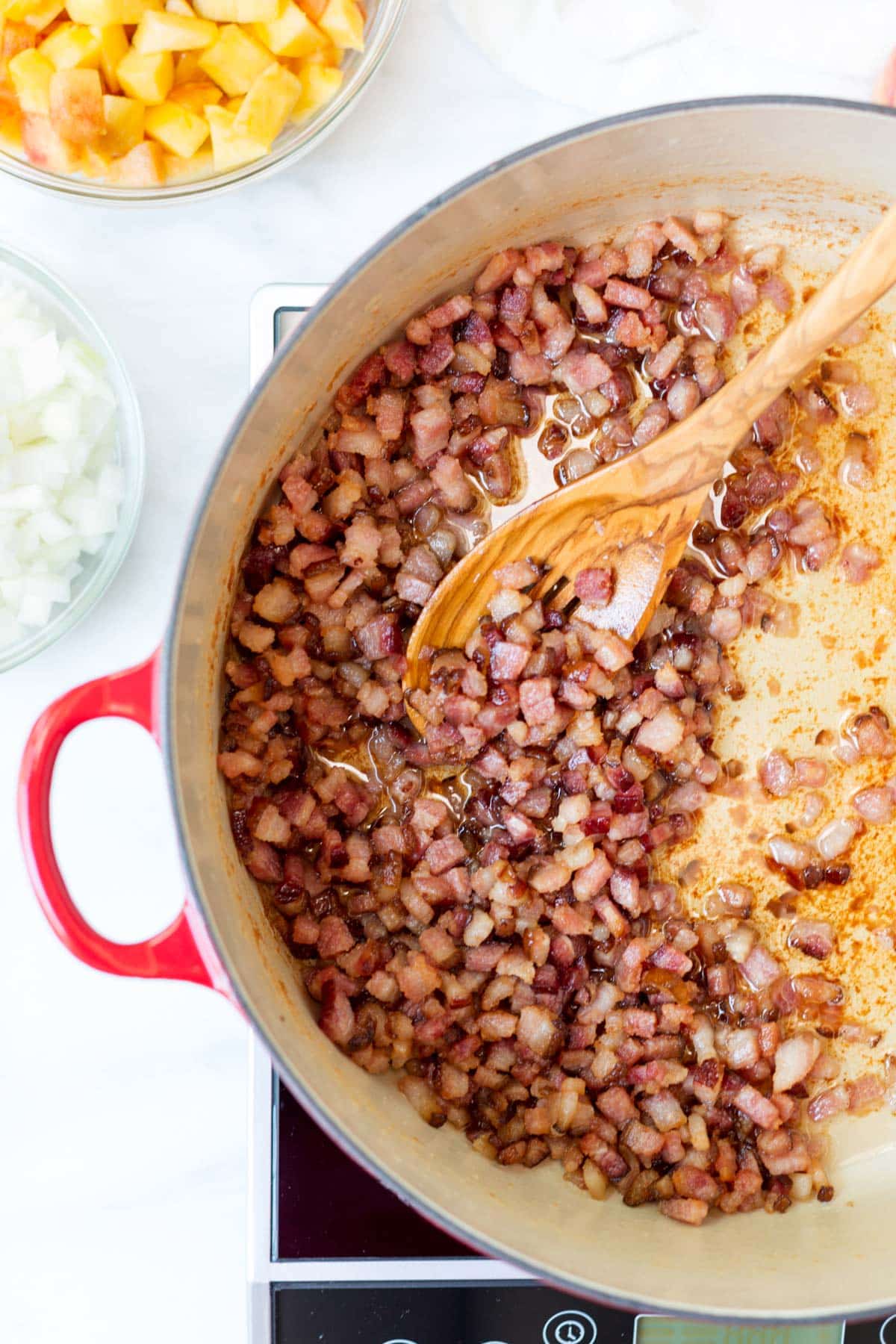 Diced bacon cooking in a Dutch oven until browned and crisp, with rendered bacon fat coating the bottom of the pot. A wooden spoon stirs the bacon while bowls of diced onion and peaches wait nearby for the next step in making peach bacon jam.