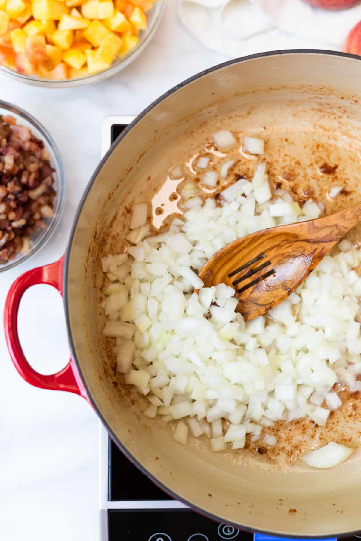 Diced onion cooking in rendered bacon fat in a Dutch oven, with a wooden spoon stirring the onions as they soften. Bowls of cooked bacon and diced peaches sit nearby, ready to be added for peach bacon jam.
