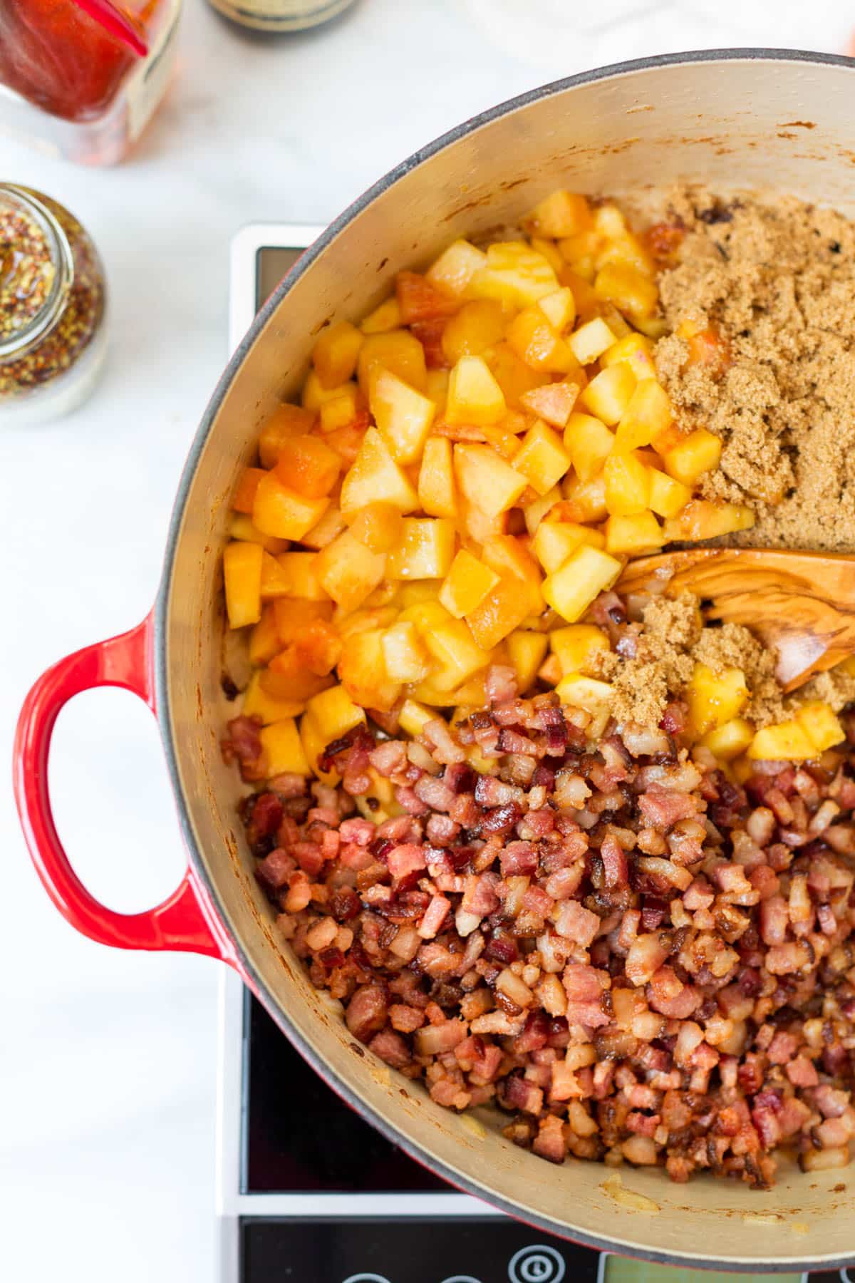Diced peaches, crispy bacon, and brown sugar added to a Dutch oven after the onions have softened. A wooden spoon begins stirring the ingredients together to make peach bacon jam.
