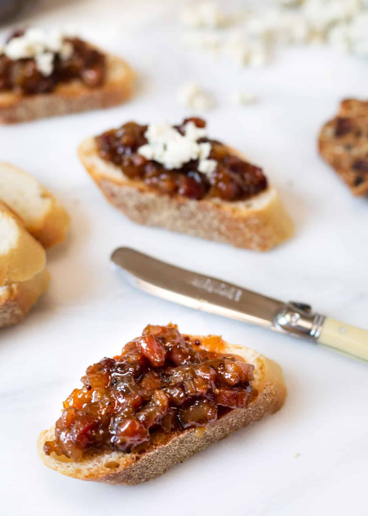 Toasted crostini topped with glossy peach bacon jam, with some pieces finished with crumbled goat cheese. The appetizers are arranged on a white serving board alongside a spreader knife, highlighting the chunky sweet-and-savory jam made with peaches, bacon, and onions.