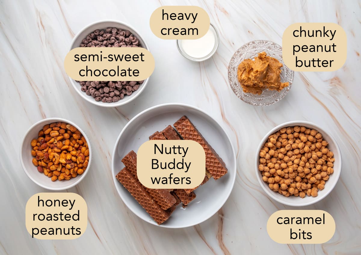 Overhead view of ingredients for chocolate peanut butter caramel bark arranged on a white marble surface, including semi-sweet chocolate chips, heavy cream, chunky peanut butter, honey roasted peanuts, caramel bits, and chocolate-covered Nutty Buddy wafers. Each ingredient is displayed in a separate bowl or plate with a beige label identifying it.
