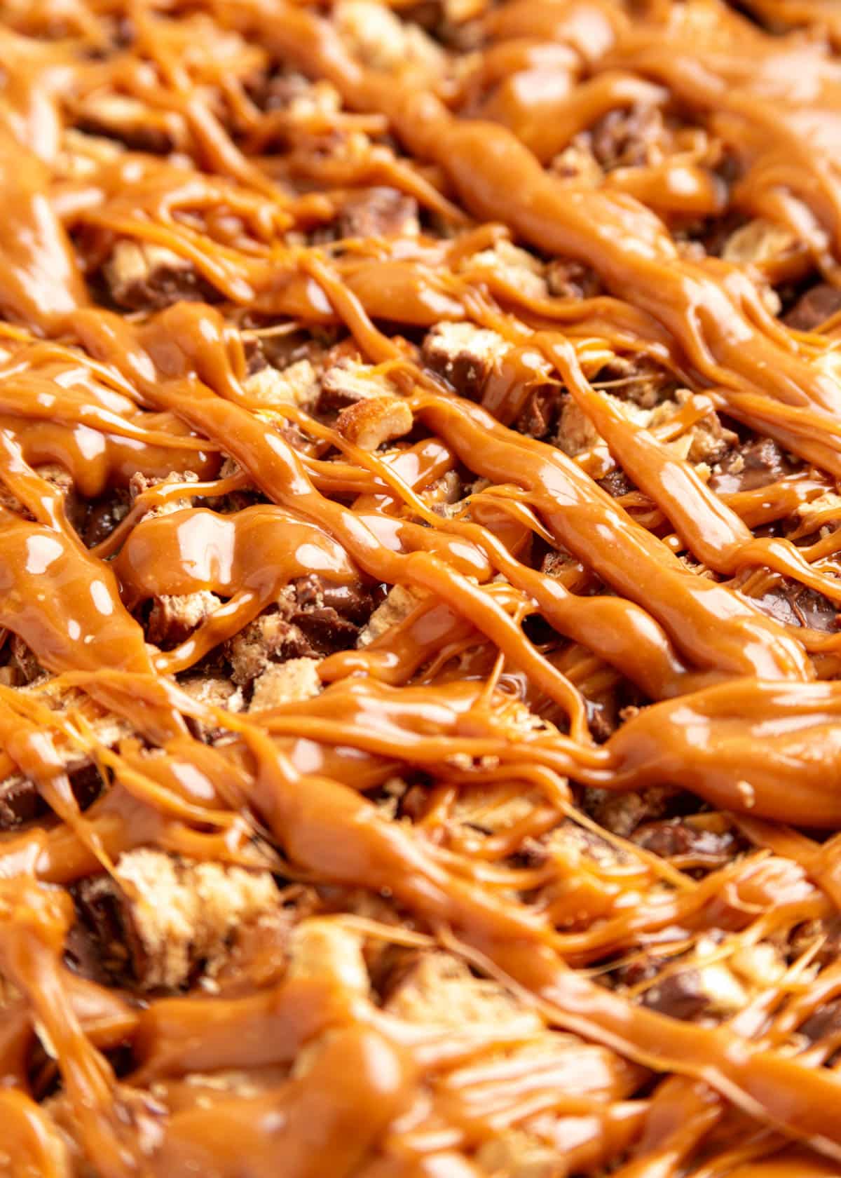 Extreme close-up of chocolate peanut butter caramel bark topped with chopped Nutty Buddy wafer cookies, crushed peanuts, and thick ribbons of glossy melted caramel drizzled across the surface.