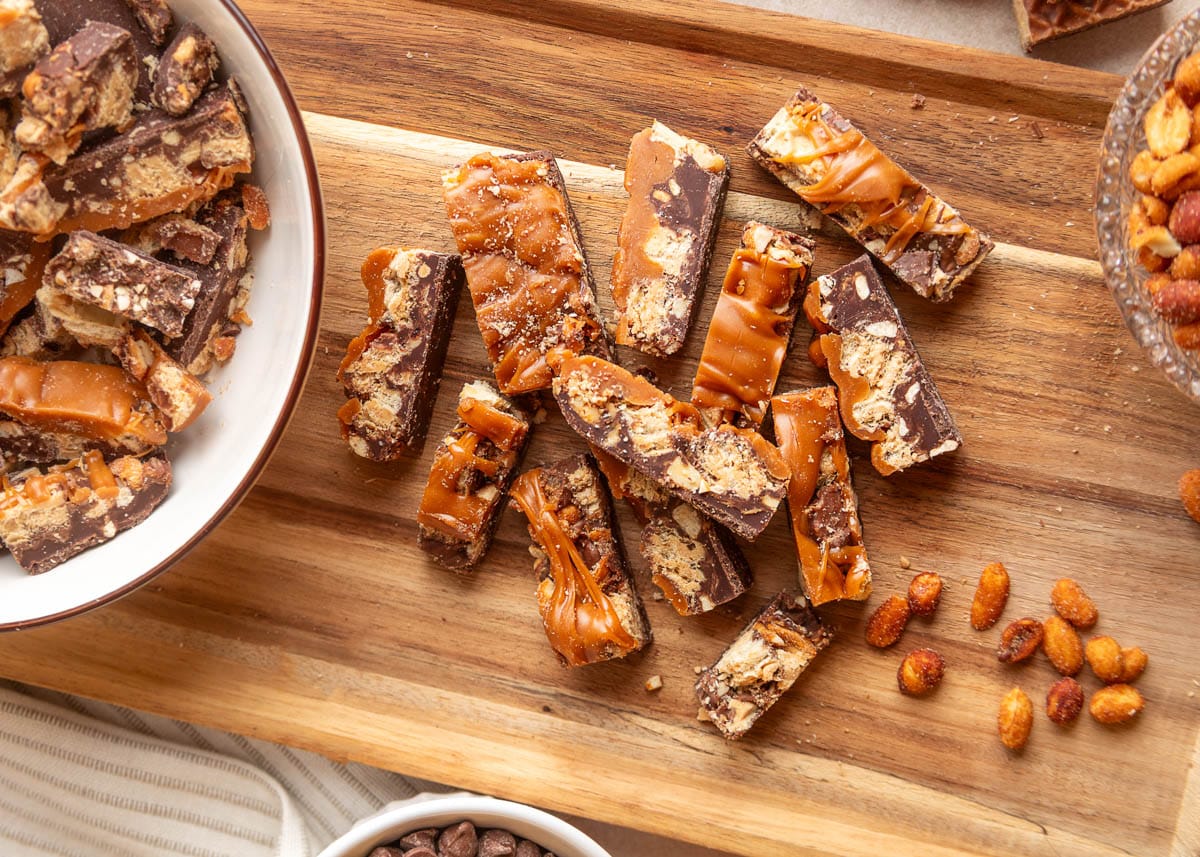 Overhead view of pieces of chocolate peanut butter caramel bark scattered across a wooden cutting board, showing layers of semi-sweet chocolate, chopped Nutty Buddy wafers, peanuts, and thick caramel drizzle. A bowl of additional bark sits on the left, with honey roasted peanuts scattered on the right.