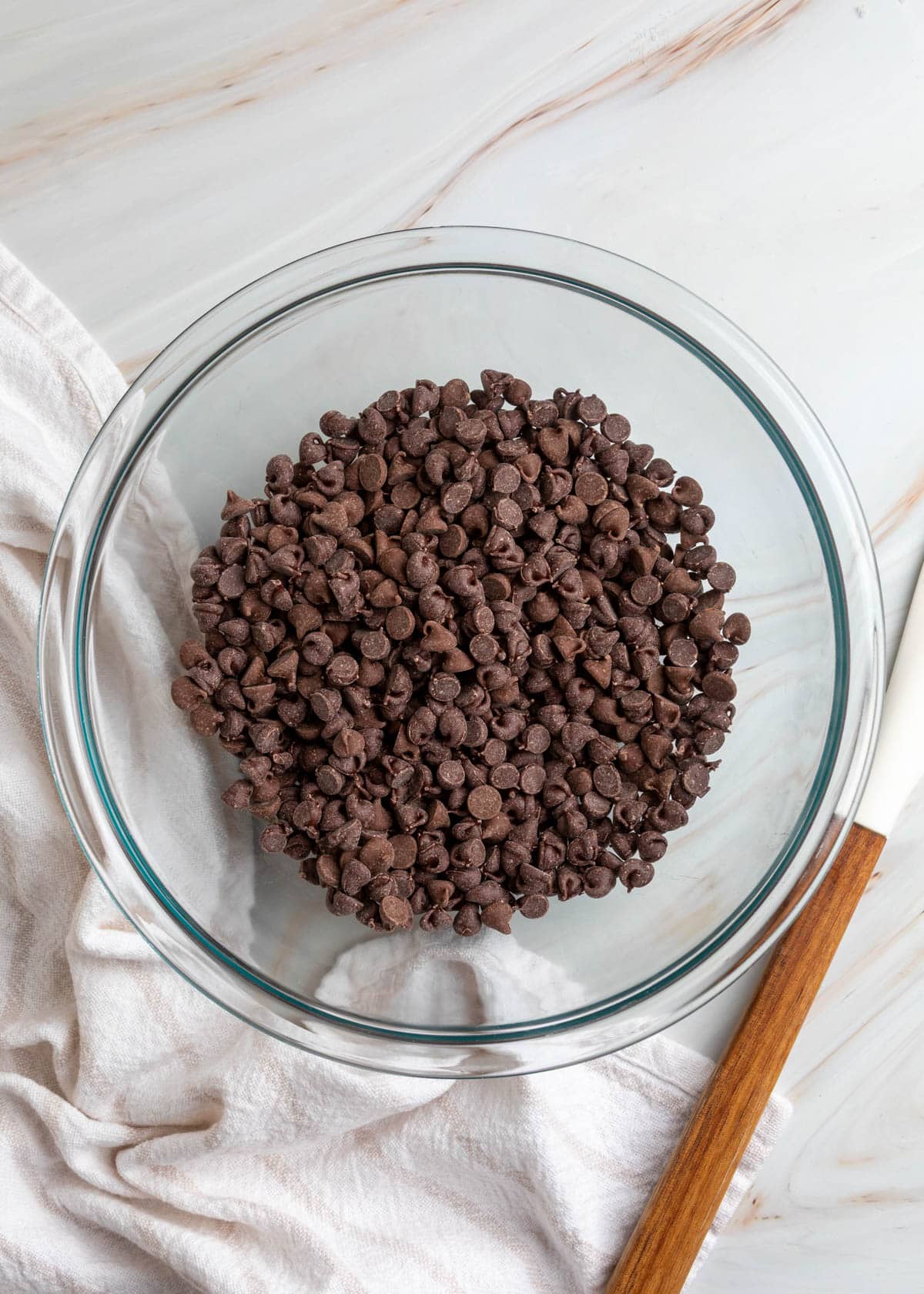 Overhead view of a clear glass mixing bowl filled with semi-sweet chocolate chips, resting on a white kitchen towel over a light marble-patterned countertop.
