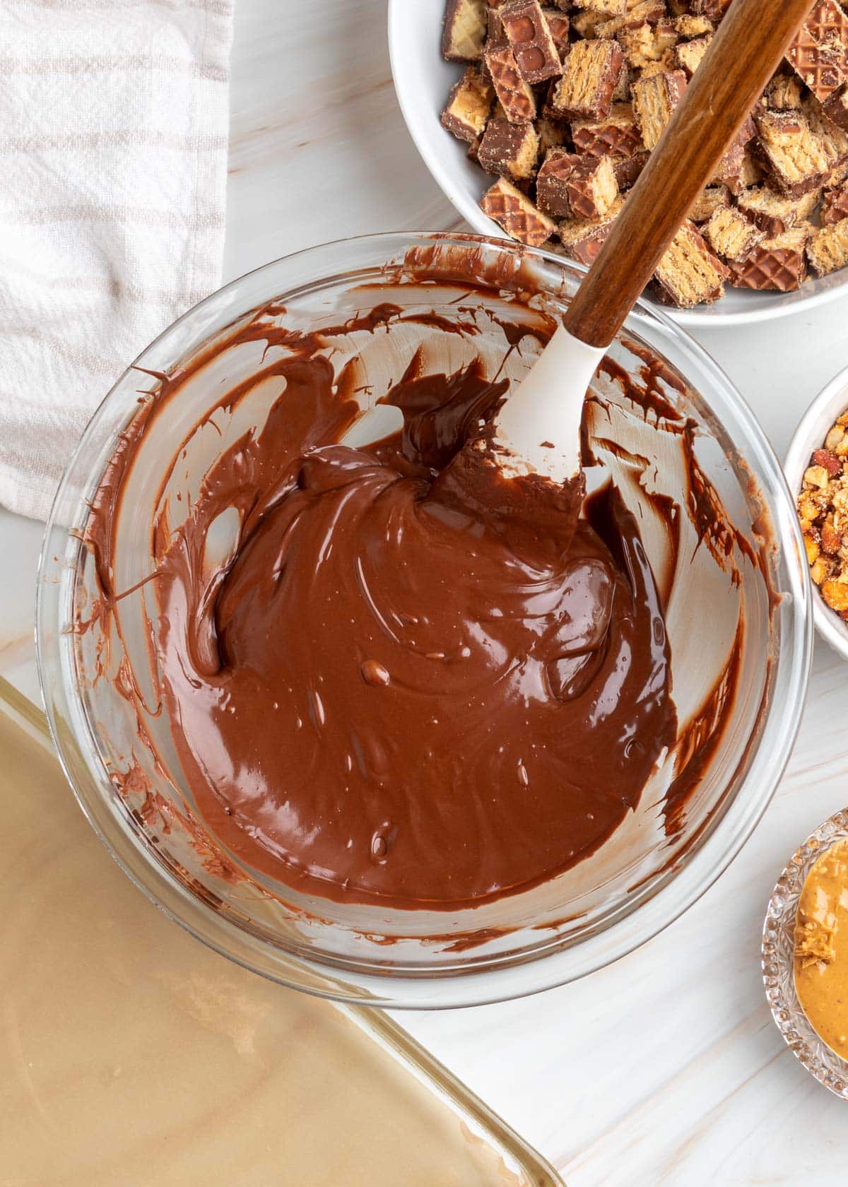 Overhead view of smooth, glossy melted chocolate in a clear glass mixing bowl with a white silicone spatula and wooden handle. Chopped Nutty Buddy wafer cookies, crushed peanuts, and peanut butter sit nearby.