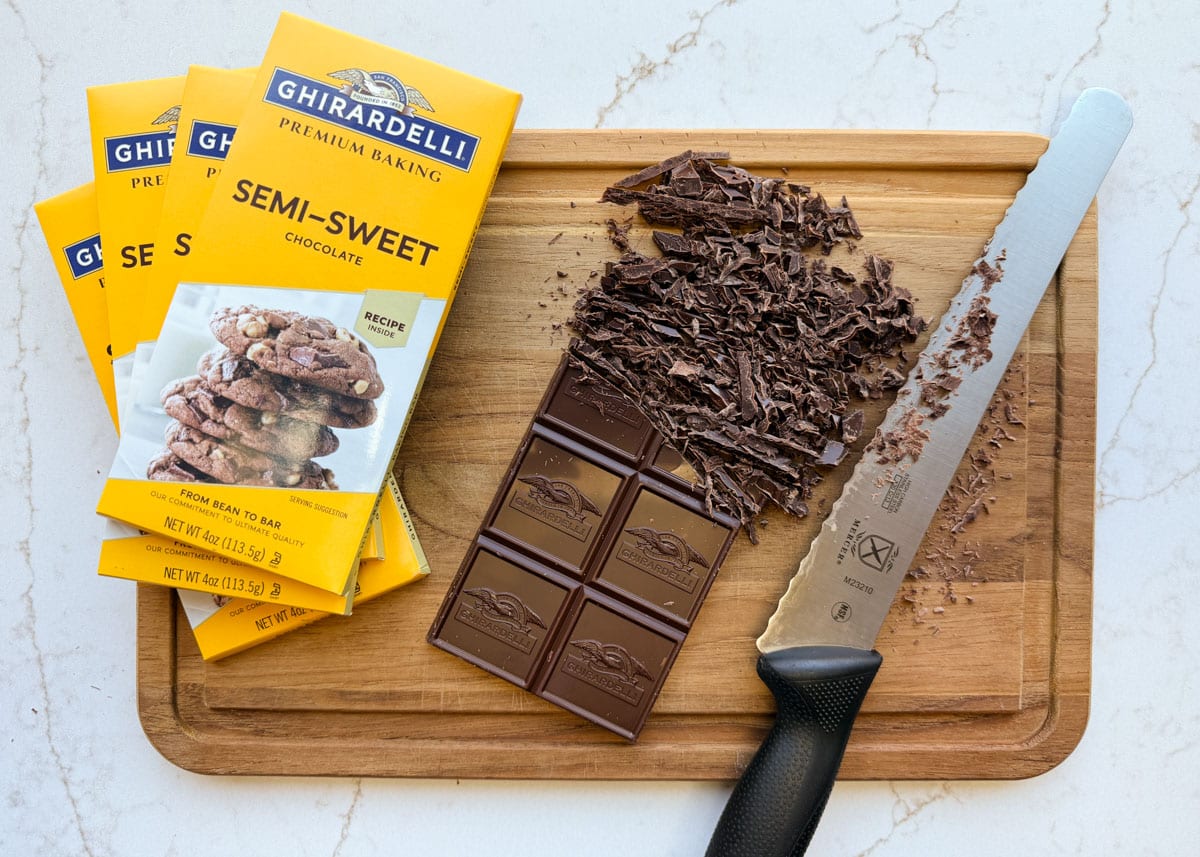 Overhead view of Ghirardelli semi-sweet baking chocolate bars on a wooden cutting board, with one partially chopped bar surrounded by chocolate shavings. A serrated knife rests beside the chopped chocolate, and several yellow Ghirardelli chocolate bar packages are stacked on the left.