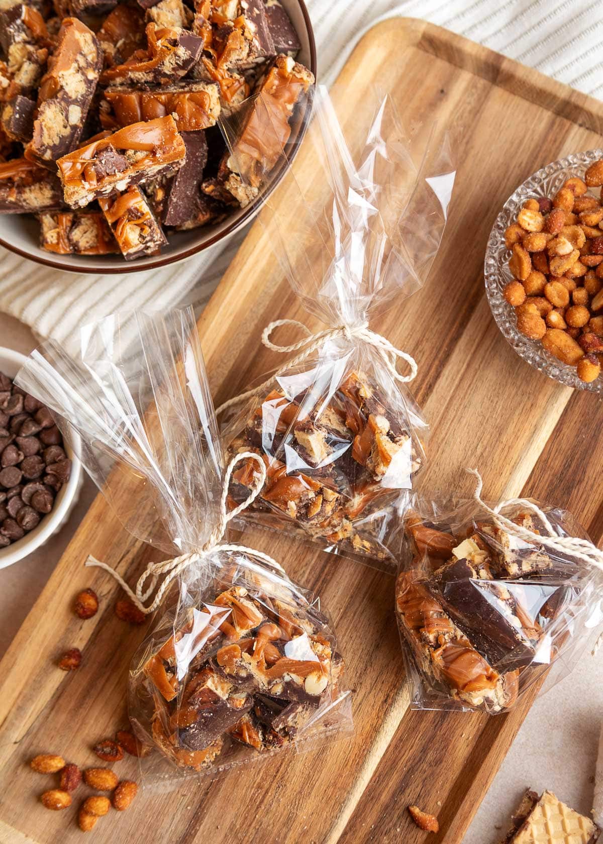 Three clear cellophane gift bags filled with pieces of chocolate peanut butter caramel bark, tied with rustic twine and arranged on a wooden cutting board. A bowl of additional bark, honey roasted peanuts, and semi-sweet chocolate chips surround the packaged treats.