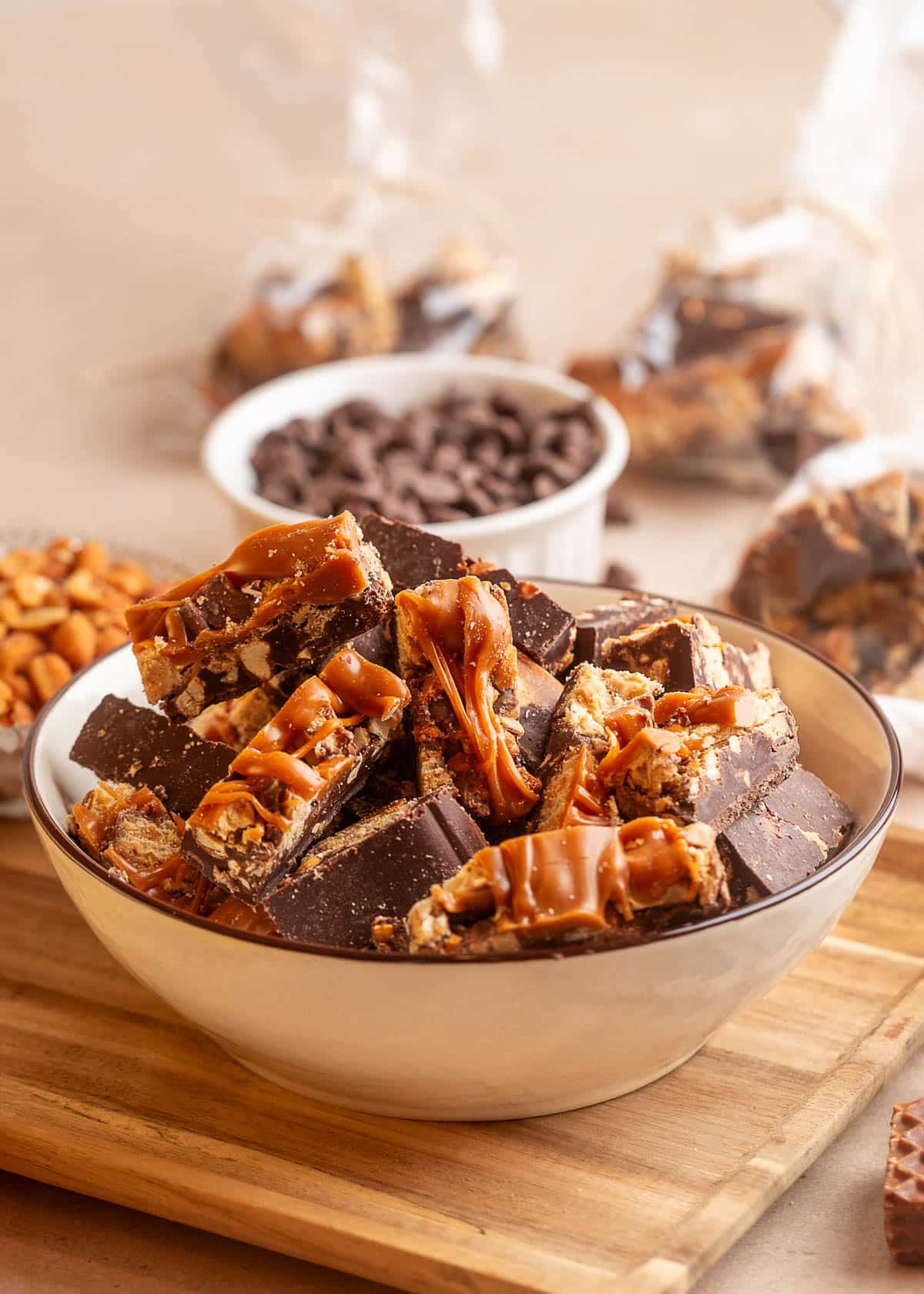A cream-colored bowl piled high with pieces of chocolate peanut butter caramel bark, featuring thick semi-sweet chocolate, crunchy Nutty Buddy wafers, peanuts, and glossy caramel drizzle. Chocolate chips, honey roasted peanuts, and cellophane gift bags filled with bark are arranged in the background.