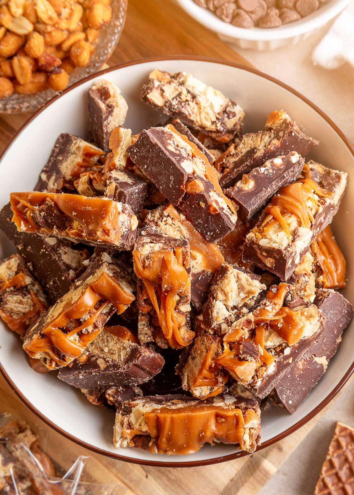 Close-up of chocolate peanut butter caramel bark broken into irregular pieces and piled in a white bowl with a brown rim. The pieces reveal layers of chocolate, crunchy peanut butter wafers, peanuts, and thick, gooey caramel stretching between the pieces, with bowls of honey roasted peanuts and caramel bits in the background.