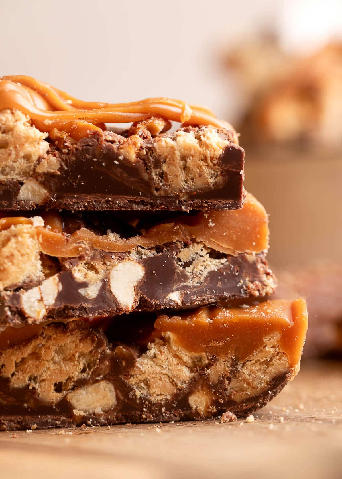 Extreme close-up of three stacked pieces of chocolate peanut butter caramel bark, highlighting thick layers of semi-sweet chocolate with embedded peanuts and crunchy wafer pieces, topped with rich caramel and glossy caramel drizzle.