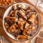 Overhead view of a cream-colored bowl filled with chocolate peanut butter caramel bark, featuring thick pieces of semi-sweet chocolate studded with peanuts and crunchy Nutty Buddy wafers, topped with generous ribbons of caramel. The bowl rests on a wooden cutting board, surrounded by honey roasted peanuts and wafer pieces.