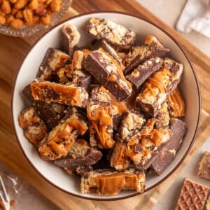 Overhead view of a cream-colored bowl filled with chocolate peanut butter caramel bark, featuring thick pieces of semi-sweet chocolate studded with peanuts and crunchy Nutty Buddy wafers, topped with generous ribbons of caramel. The bowl rests on a wooden cutting board, surrounded by honey roasted peanuts and wafer pieces.