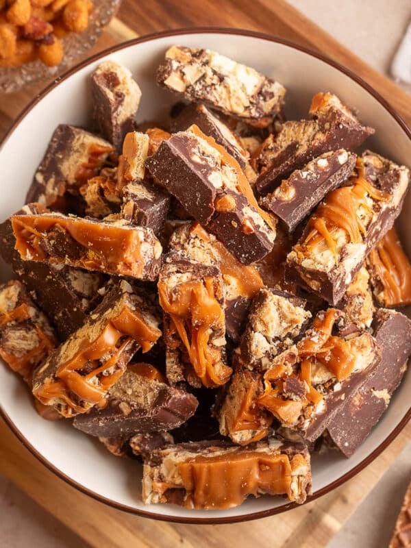 Overhead view of a cream-colored bowl filled with chocolate peanut butter caramel bark, featuring thick pieces of semi-sweet chocolate studded with peanuts and crunchy Nutty Buddy wafers, topped with generous ribbons of caramel. The bowl rests on a wooden cutting board, surrounded by honey roasted peanuts and wafer pieces.