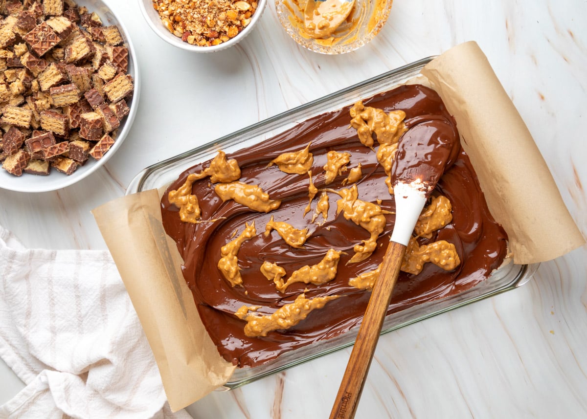Overhead view of melted chocolate spread in a parchment-lined glass baking dish, with dollops of chunky peanut butter scattered across the surface. A chocolate-coated spatula rests on top, with bowls of chopped Nutty Buddy wafers, crushed honey roasted peanuts, and peanut butter nearby.