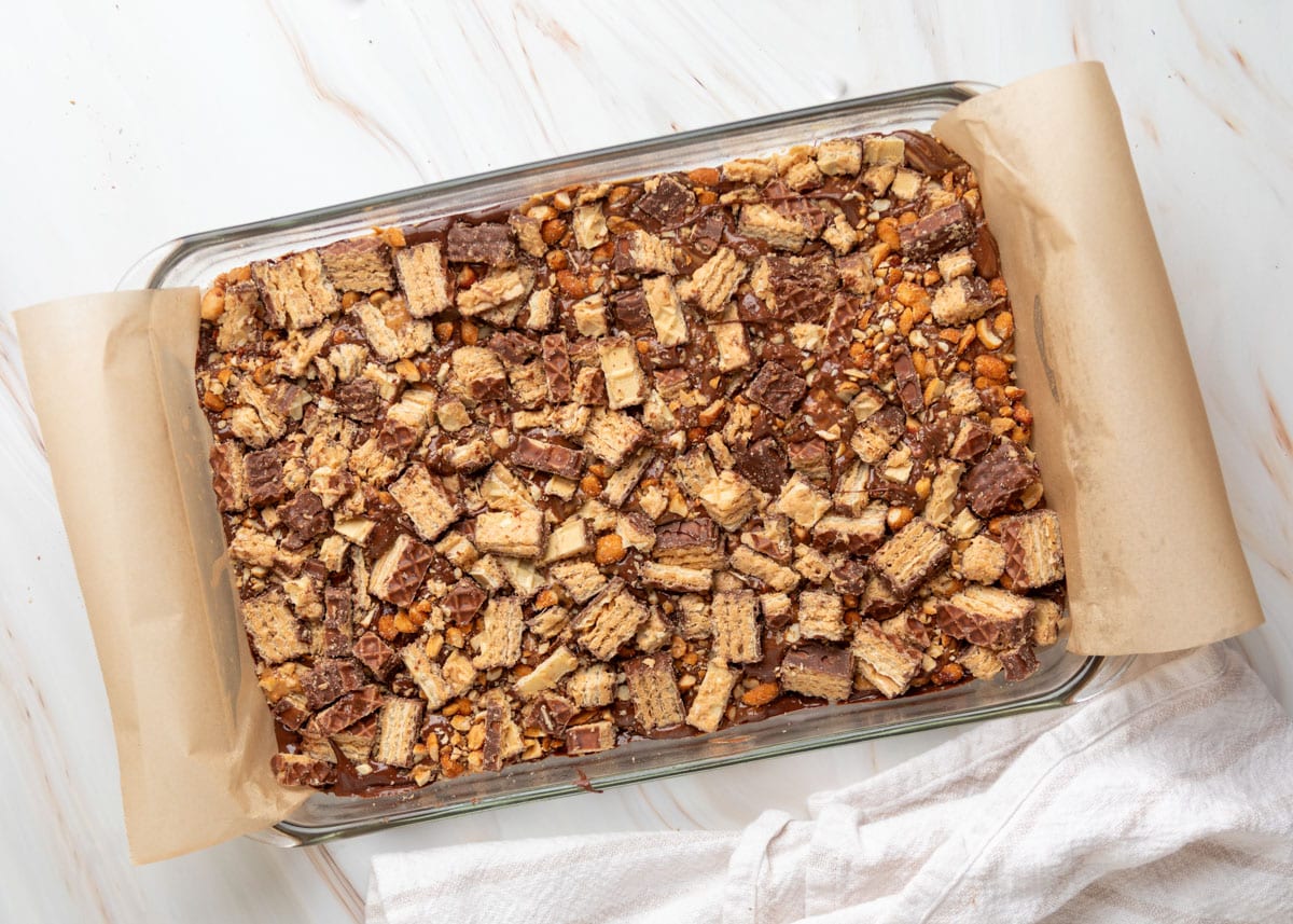 Overhead view of chocolate peanut butter caramel bark in a parchment-lined glass baking dish, generously topped with chopped chocolate-covered Nutty Buddy wafers and crushed honey roasted peanuts over a layer of melted chocolate.