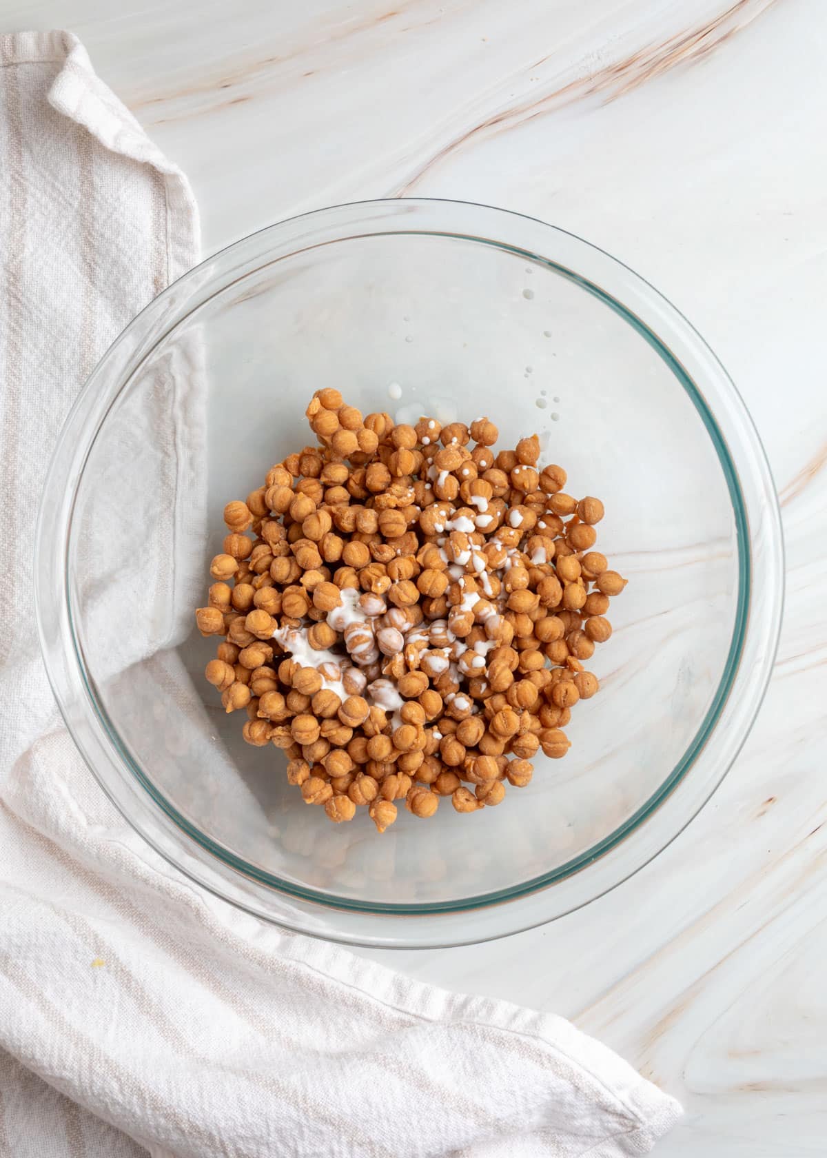 Overhead view of caramel bits in a clear glass mixing bowl with a small amount of heavy cream drizzled over them, ready to be melted into caramel sauce.