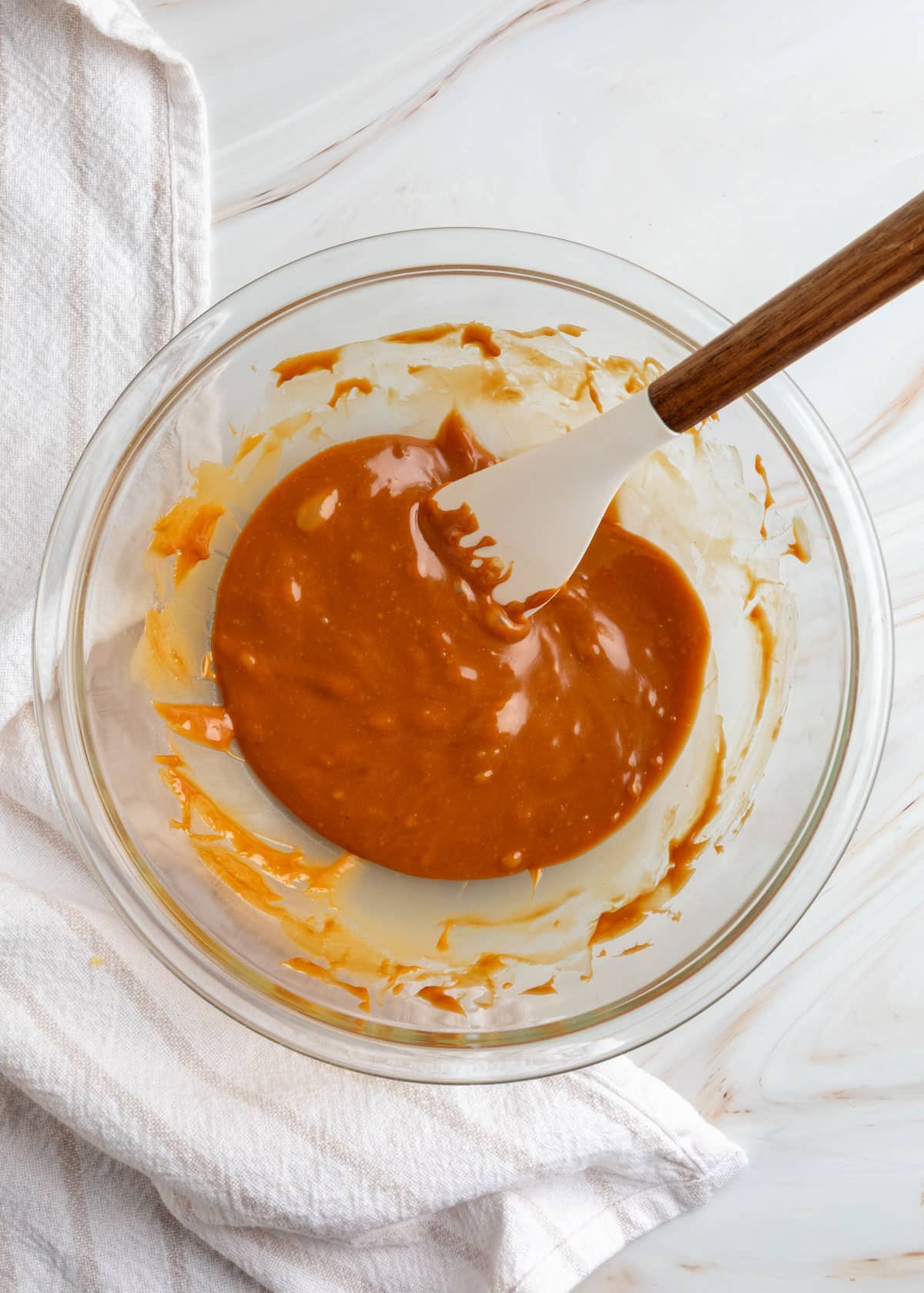 Overhead view of smooth, glossy melted caramel sauce in a clear glass mixing bowl, with a white silicone spatula resting inside and caramel coating the sides of the bowl.