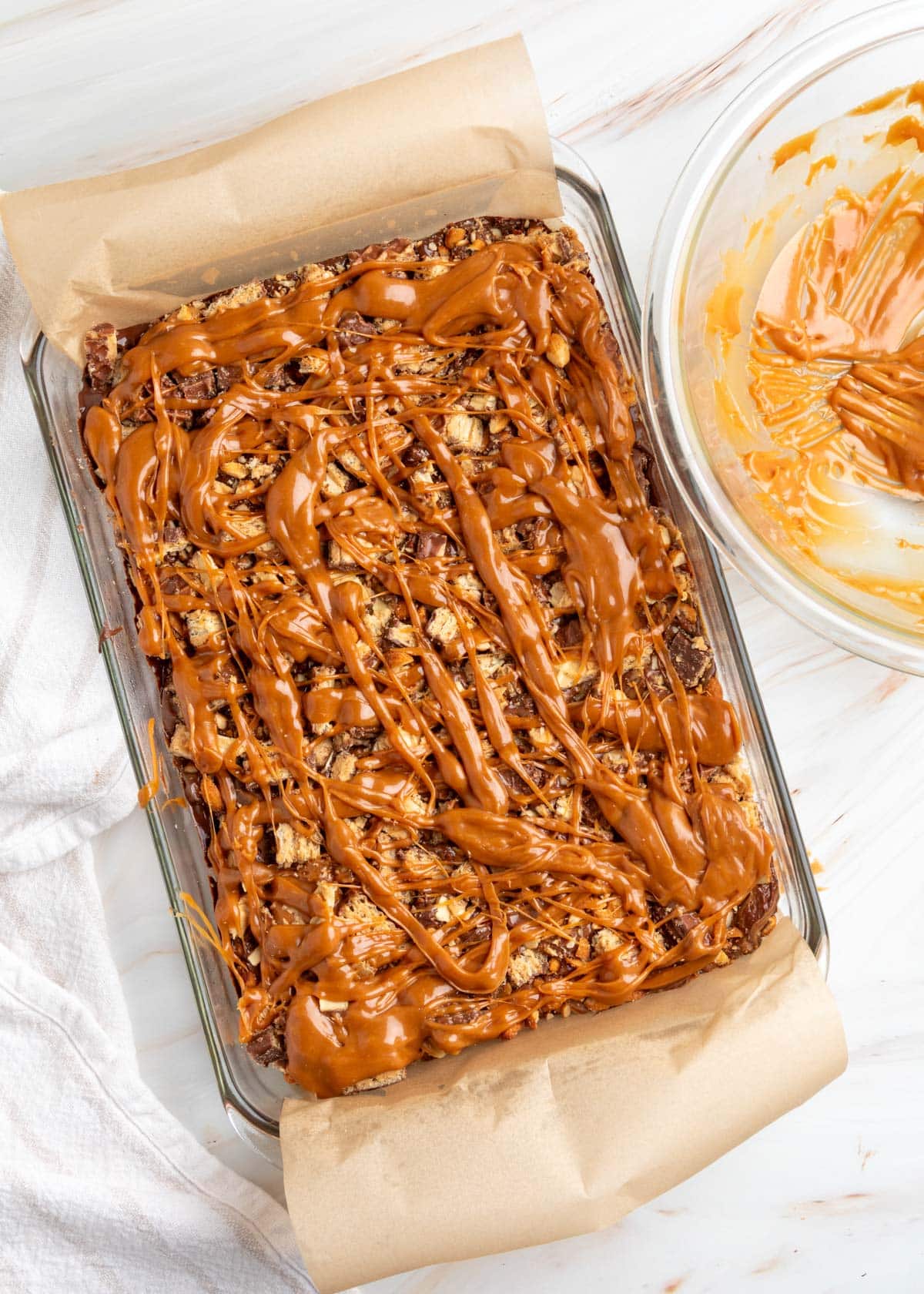 Overhead view of chocolate peanut butter caramel bark in a parchment-lined glass baking dish, covered with chopped Nutty Buddy wafers, crushed peanuts, and generous drizzles of melted caramel. A glass bowl with remaining caramel sits beside the dish.