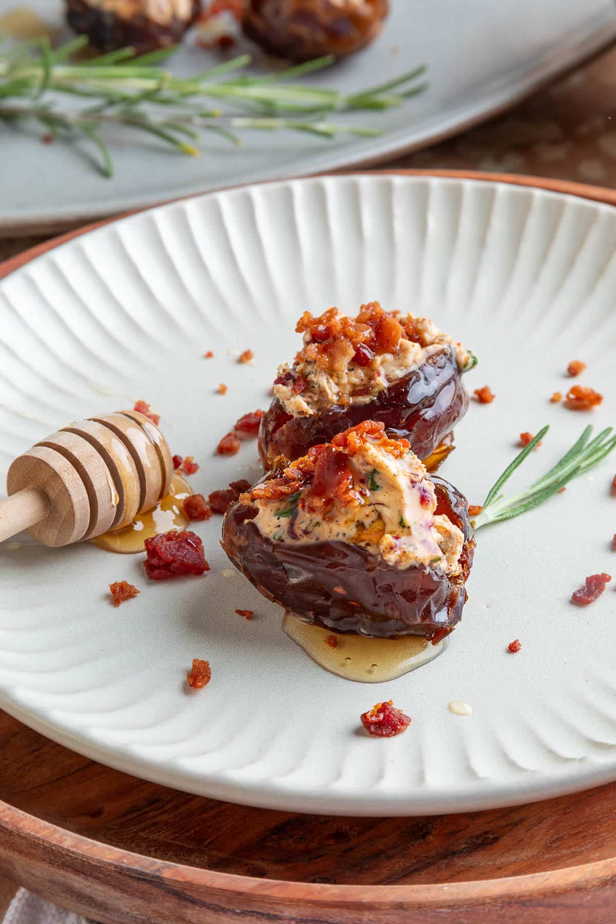 Two goat cheese stuffed Medjool dates topped with crispy bacon and hot honey are served on a plate, garnished with fresh rosemary and scattered bacon crumbles, with a honey dipper resting alongside.