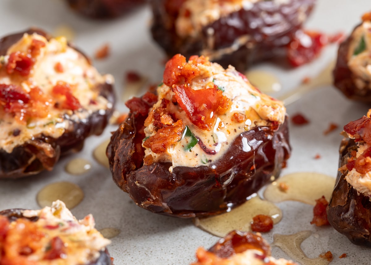 Close-up of a goat cheese stuffed Medjool date topped with crispy bacon and drizzled with hot honey, with additional stuffed dates softly blurred in the background.