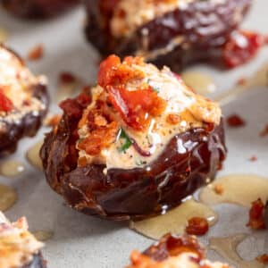 Close-up of a goat cheese stuffed Medjool date topped with crispy bacon and drizzled with hot honey, with additional stuffed dates softly blurred in the background.