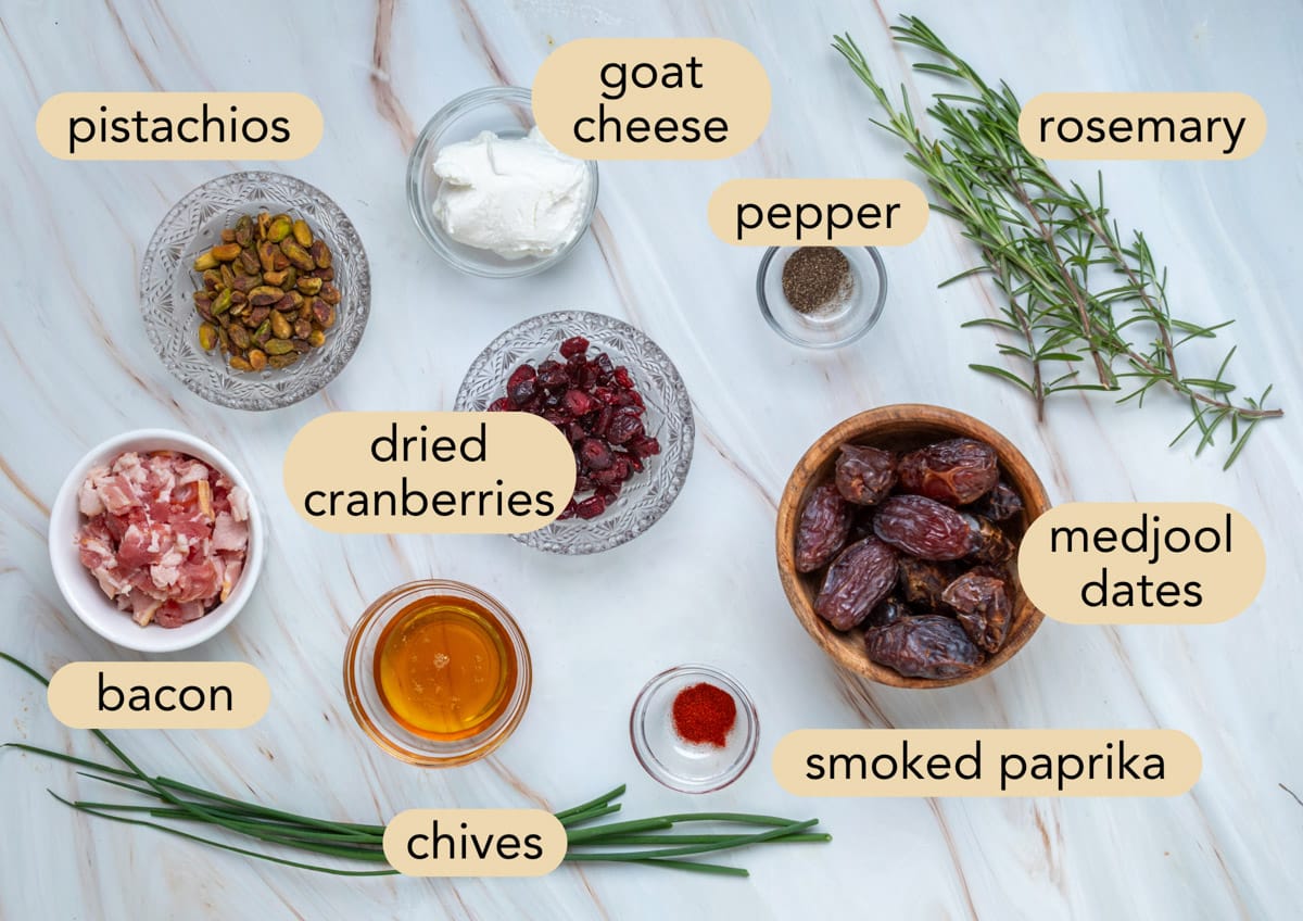 Fresh ingredients for goat cheese stuffed dates arranged on a marble surface, including Medjool dates, goat cheese, bacon, pistachios, dried cranberries, fresh rosemary, chives, smoked paprika, black pepper, and hot honey, with each ingredient labeled.