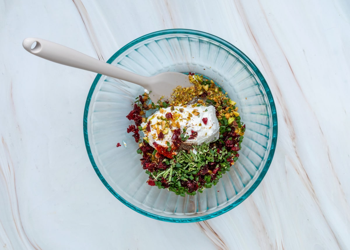 Goat cheese, chopped pistachios, dried cranberries, chives, rosemary, smoked paprika, and black pepper layered together in a glass bowl before being mixed.