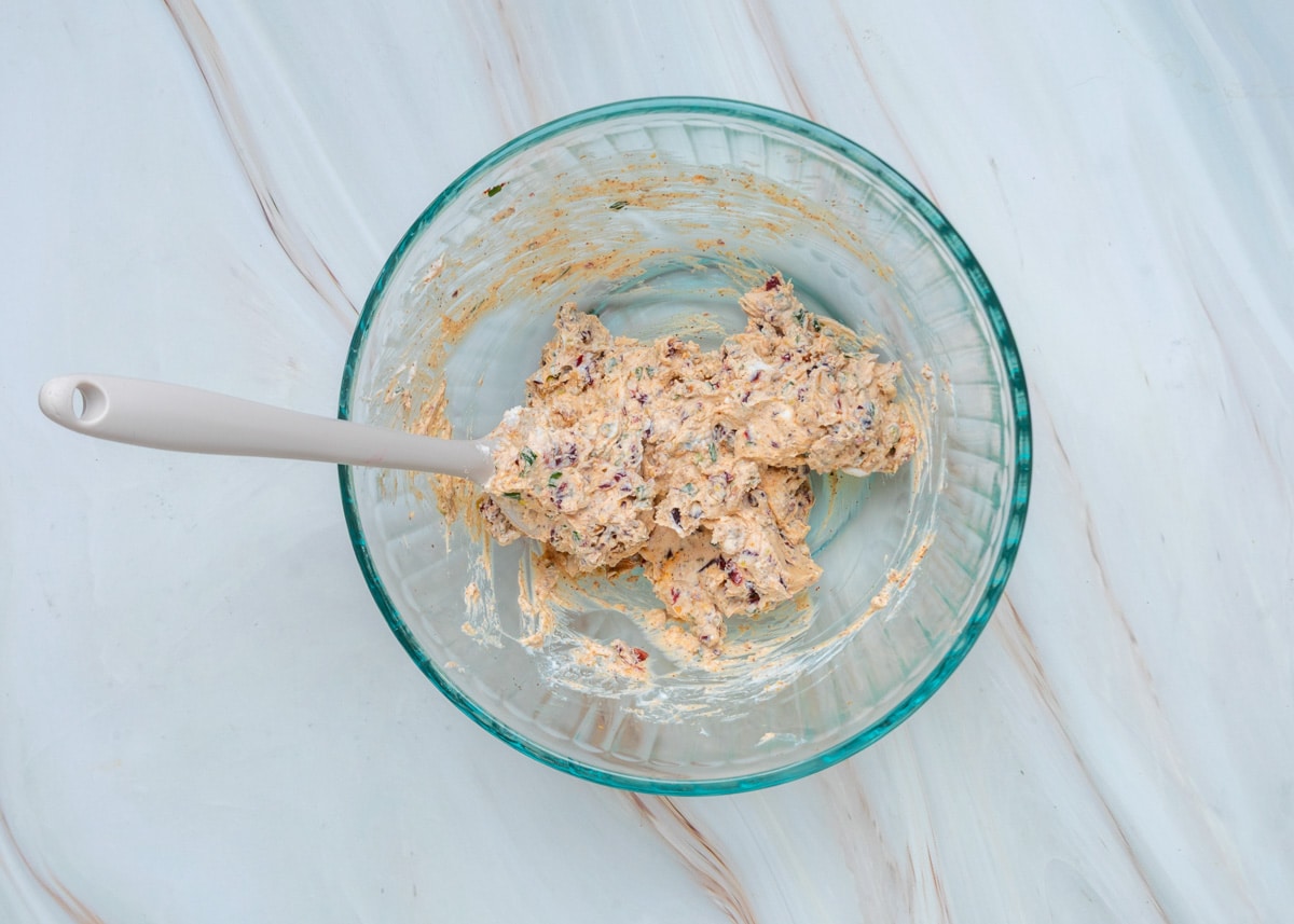 Goat cheese filling after mixing, with chopped dried cranberries, pistachios, chives, rosemary, smoked paprika, and black pepper evenly blended into a creamy mixture in a glass bowl.