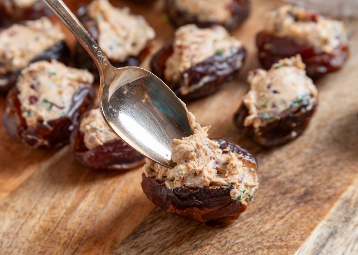 A spoon spreads the creamy goat cheese filling into split Medjool dates on a wooden board, with additional stuffed dates in the background ready for topping.