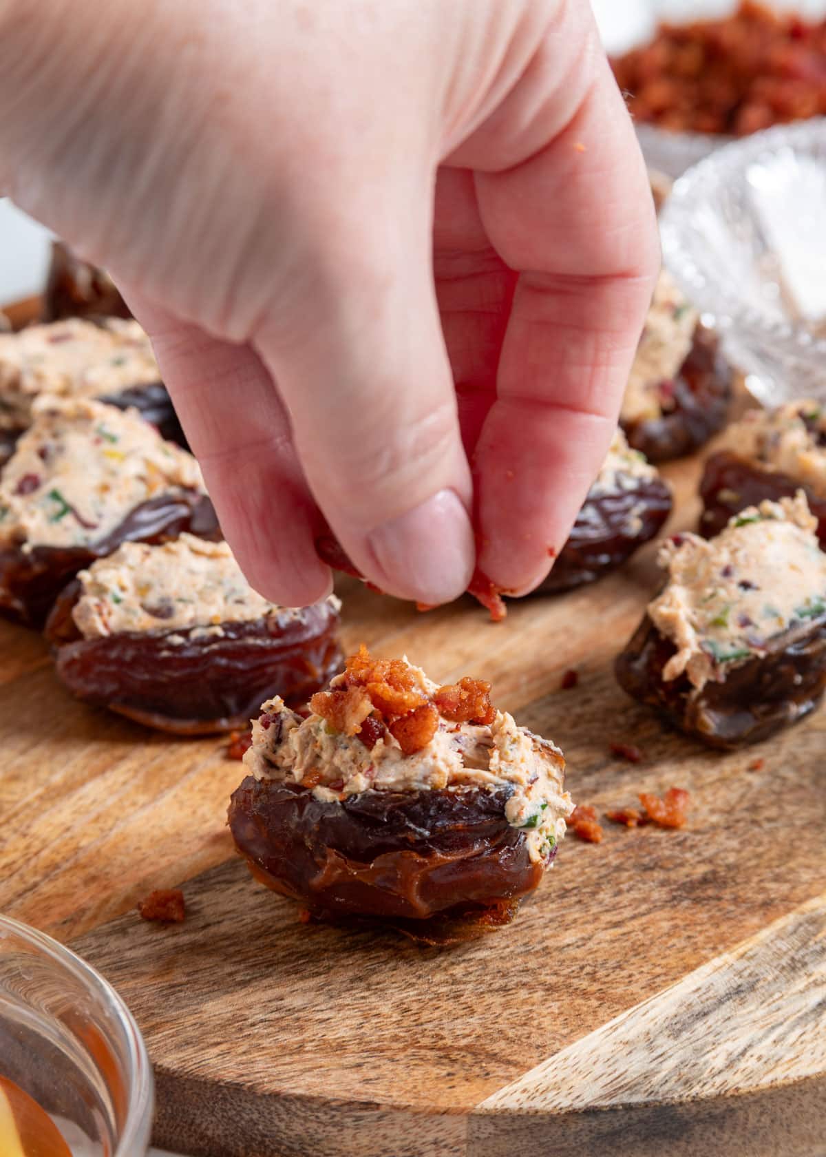 A hand sprinkles crispy bacon over a goat cheese stuffed Medjool date on a wooden serving board, finishing the appetizer before serving.