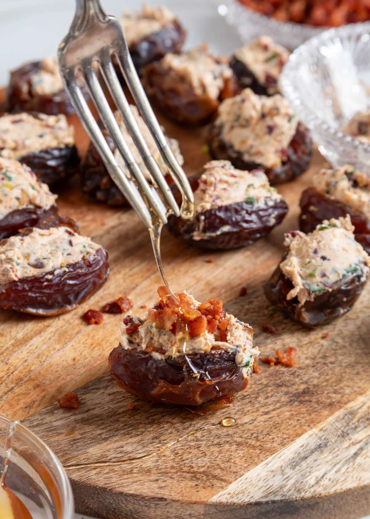 A fork drizzles hot honey over a goat cheese stuffed Medjool date topped with crispy bacon, while additional stuffed dates rest on a wooden serving board in the background.