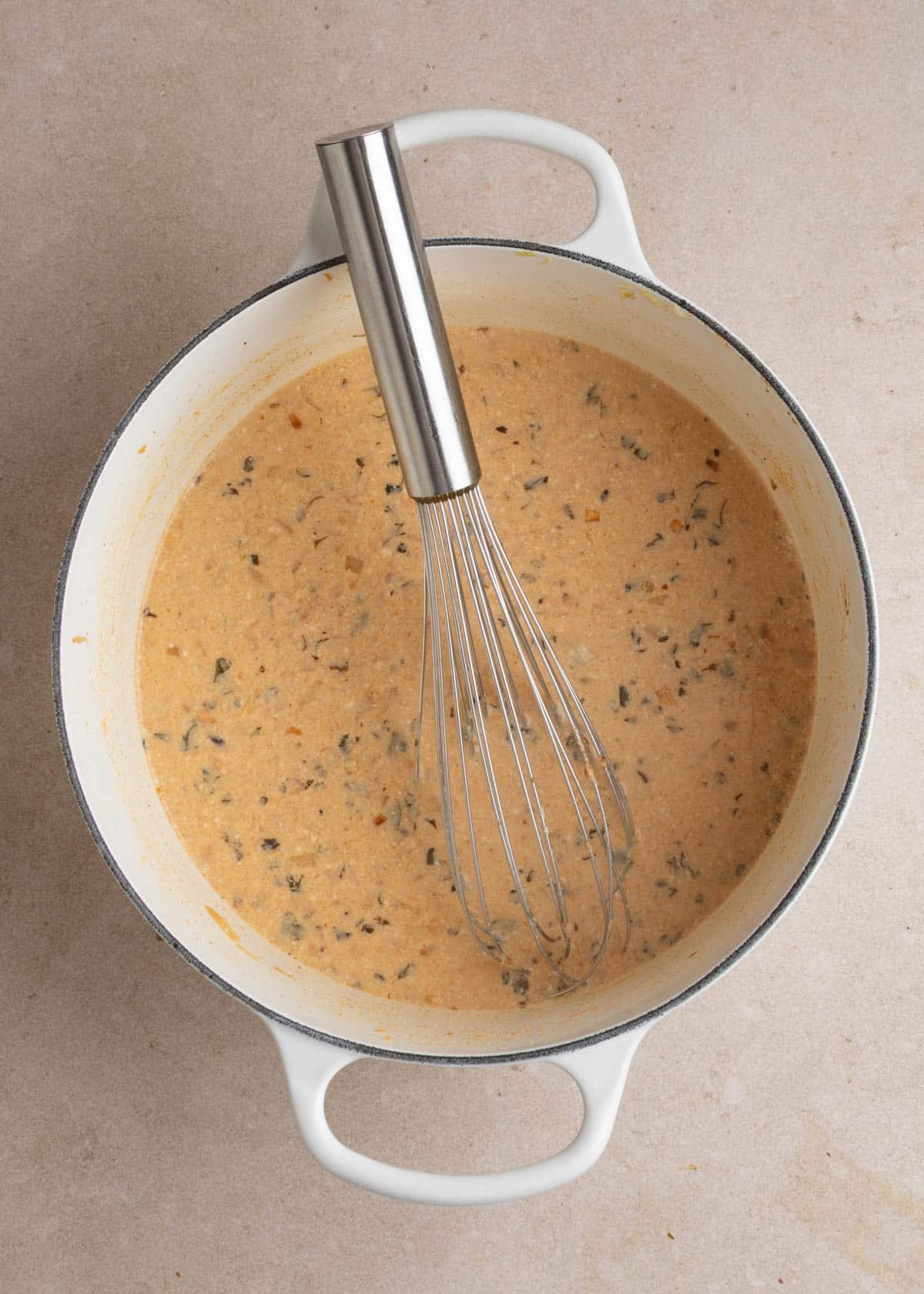 Smooth, light orange broth with flecks of herbs, spices, and diced onions in a white enameled Dutch oven, with a stainless steel whisk resting in the mixture.