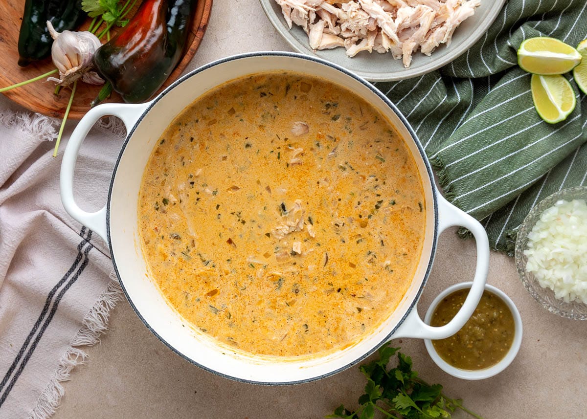 Creamy poblano white chicken chili simmered in a white enameled Dutch oven with diced roasted poblanos, salsa verde, cannellini beans, onions, and spices stirred into the broth, before the shredded chicken and fresh cilantro are added.