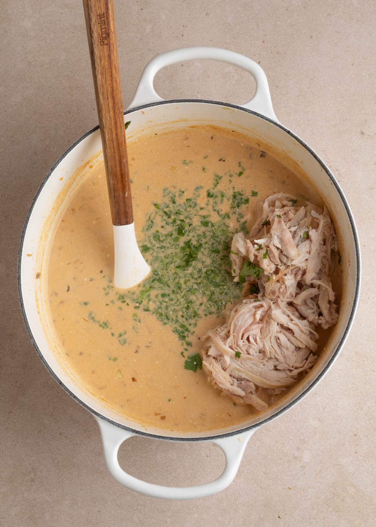 Shredded rotisserie chicken and freshly chopped cilantro added to creamy poblano white chicken chili in a white enameled Dutch oven, with a silicone spatula ready to stir everything together.