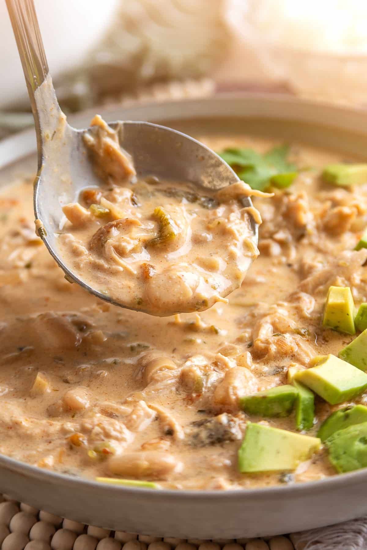 Close-up of a metal spoon lifting a hearty scoop of creamy poblano white chicken chili, showing tender shredded chicken, cannellini beans, and diced poblano peppers in a thick, lightly orange-tinted broth. Diced avocado garnishes the bowl below.