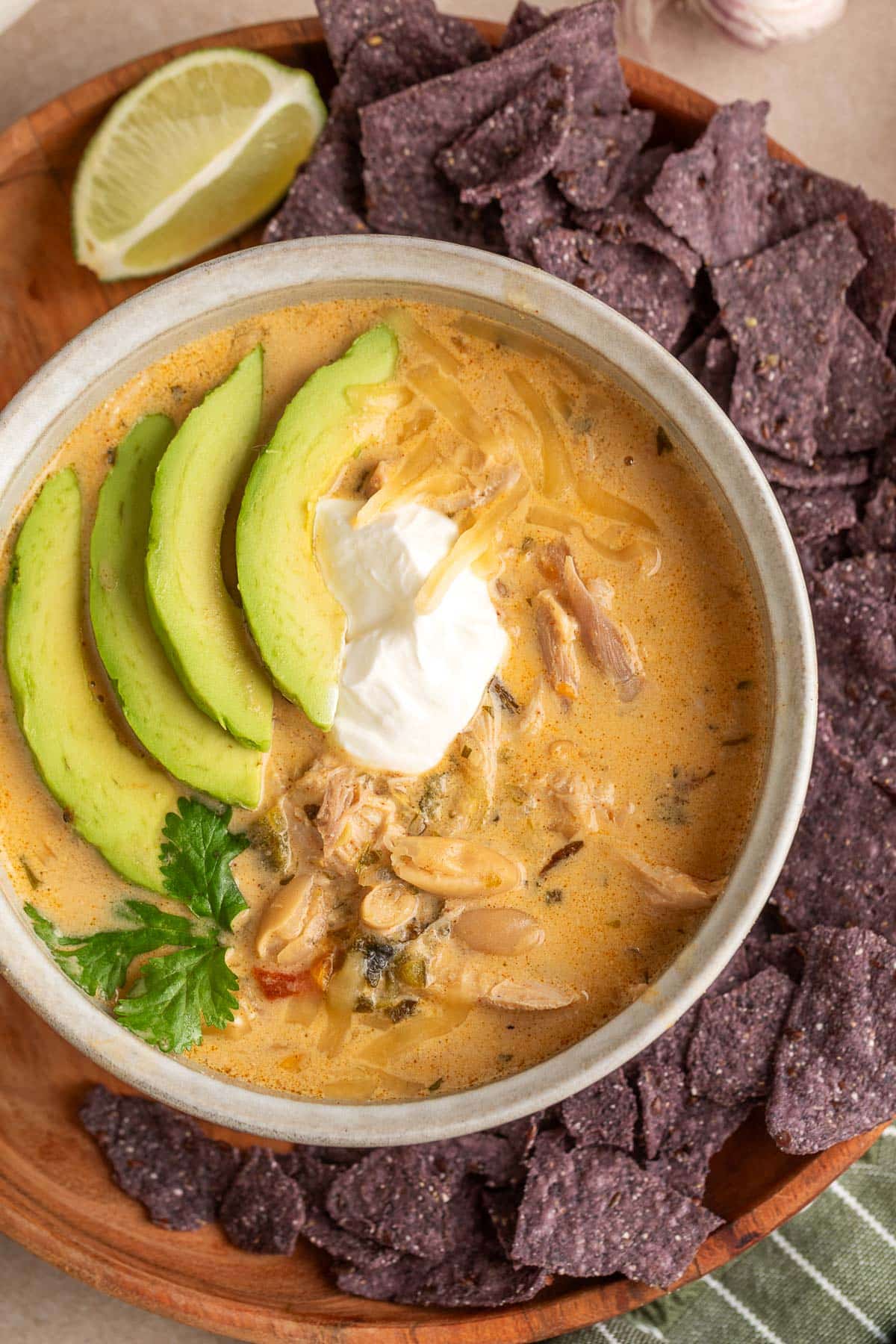 Creamy poblano white chicken chili topped with avocado slices, sour cream, shredded cheese, and cilantro, served with blue corn tortilla chips and a lime wedge on a wooden plate.