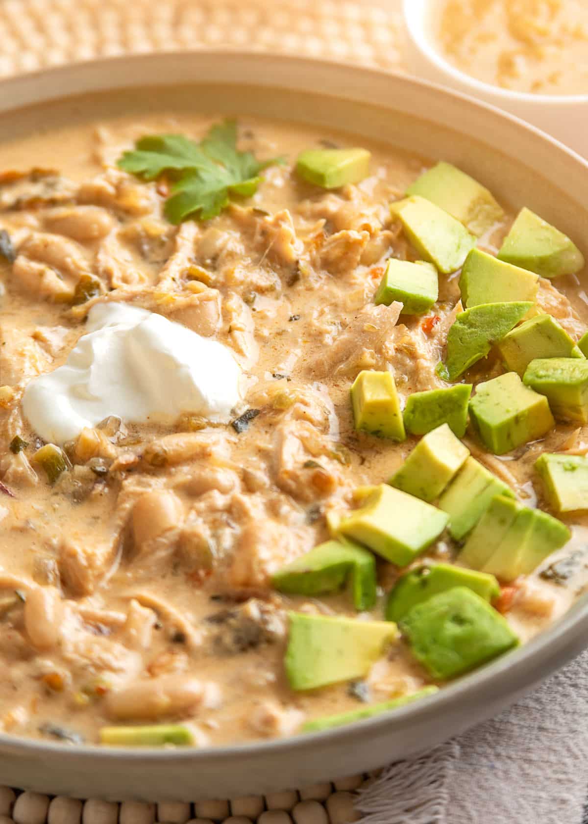 Close-up of creamy poblano white chicken chili in a shallow ceramic bowl, filled with shredded chicken, cannellini beans, and diced poblano peppers in a thick, golden-orange broth. Topped with diced avocado, a dollop of sour cream, and fresh cilantro, with a small bowl of shredded cheese in the background.