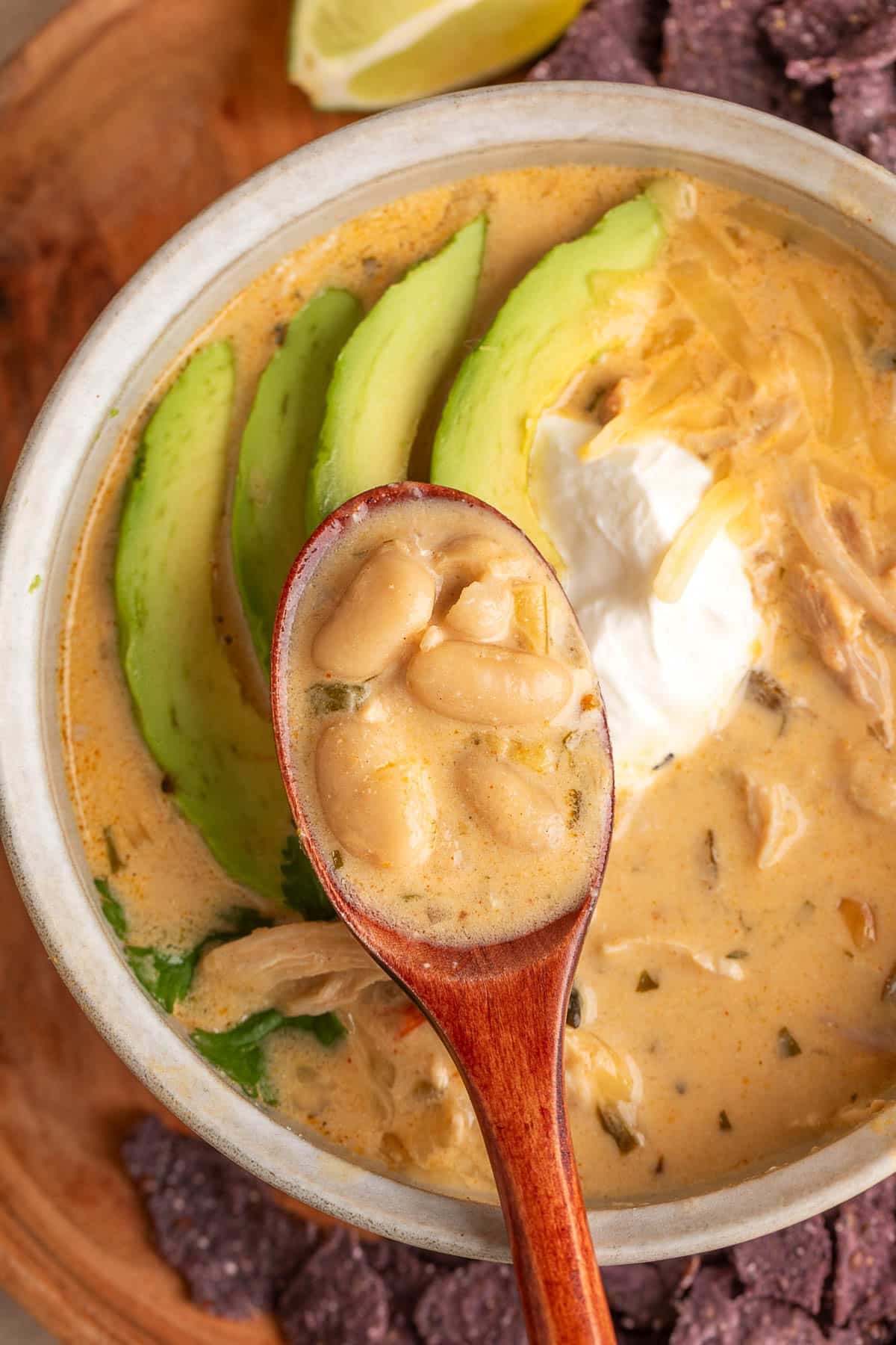Close-up of creamy poblano white chicken chili in a rustic ceramic bowl, with a wooden spoon holding plump cannellini beans in a golden-orange broth. The chili is topped with sliced avocado, a dollop of sour cream, and shredded cheese, with tender chicken, cilantro, blue corn tortilla chips, and a lime wedge visible around the bowl.