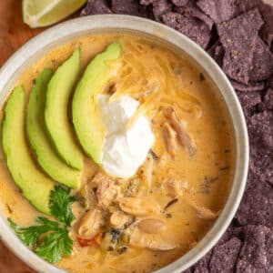 Overhead close-up of creamy poblano white chicken chili in a rustic ceramic bowl, filled with shredded chicken, cannellini beans, and diced poblano peppers in a golden-orange broth. Topped with sliced avocado, a dollop of sour cream, shredded cheese, and fresh cilantro, with blue corn tortilla chips and a lime wedge alongside the bowl.