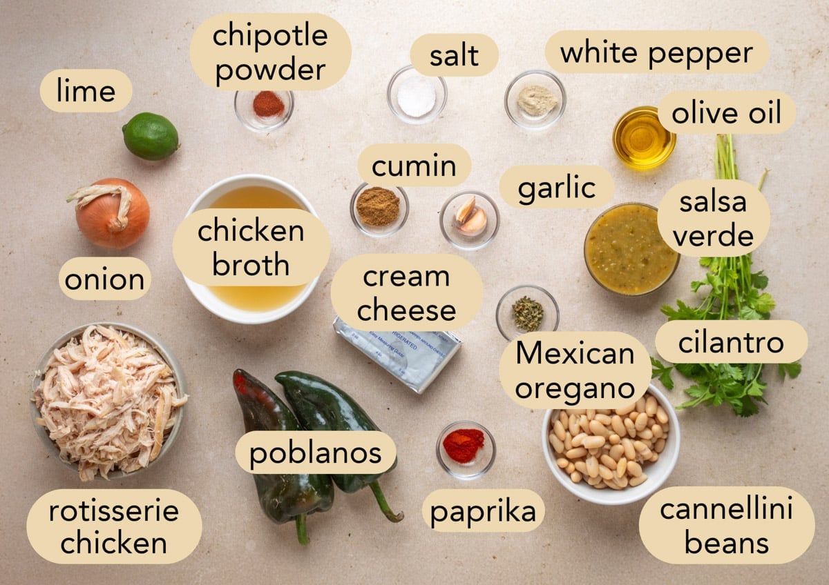 Overhead view of the ingredients for poblano white chicken chili, including rotisserie chicken, poblano peppers, cannellini beans, tomatillo salsa, cream cheese, chicken broth, onion, garlic, cilantro, lime, olive oil, and seasonings labeled for the recipe.