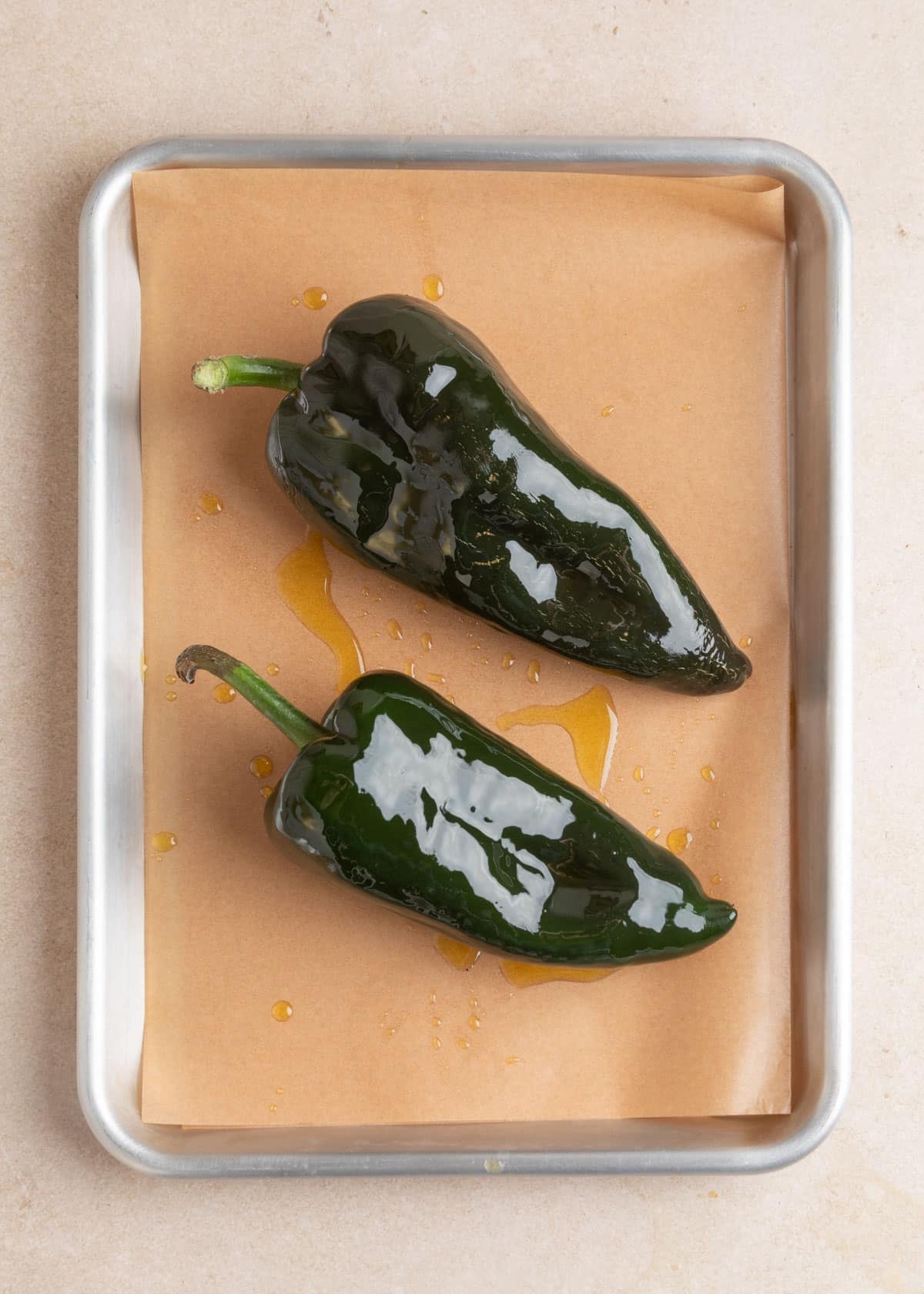 Two whole, glossy dark green poblano peppers coated with olive oil rest on a parchment-lined metal baking sheet, ready to be roasted for poblano white chicken chili.
